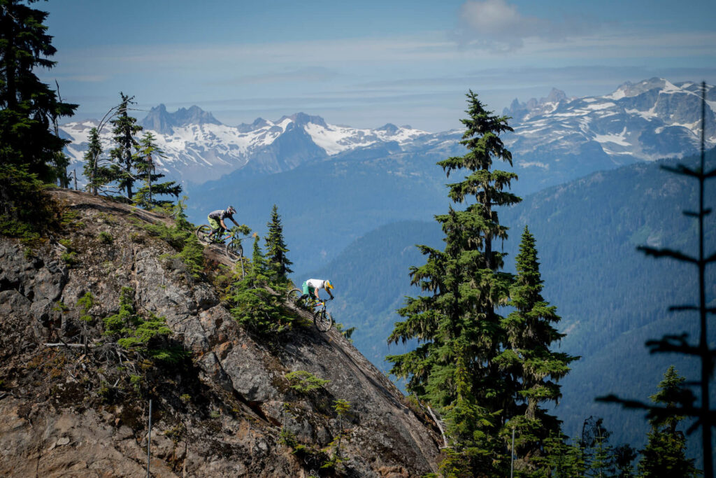 Two bikers biking down rocky trail with snow-covered mountains behind them 